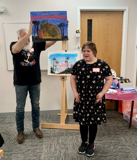 An artist and an instructor holding up a finished painting at the Joyful Arts art show