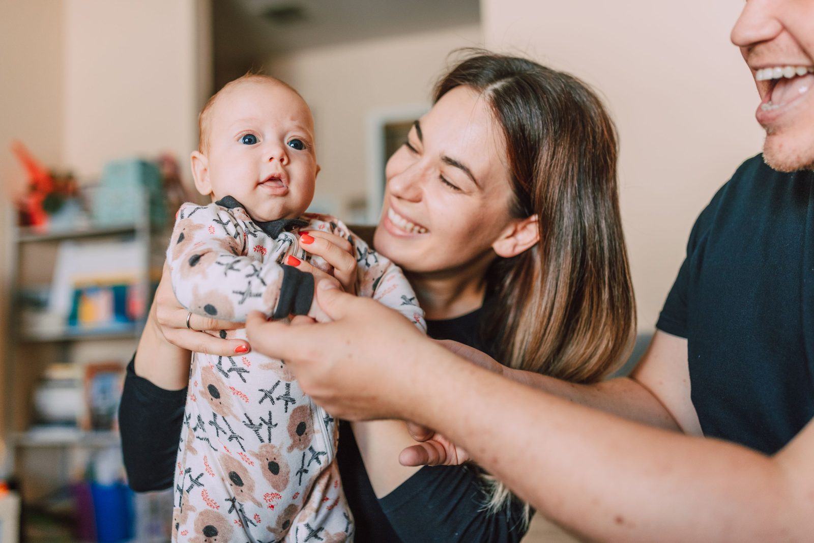 Una madre sonriente sostiene en alto a su bebé, quien mira hacia las manos extendidas de otro adulto